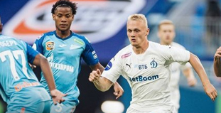 На изображении несколько футболистов в борьбе за мяч на поле. Один играет в белой форме команды Динамо, другие в бирюзовой форме команды Зенит. На заднем плане размыт логотип или баннер