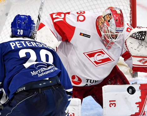 На изображении хоккейный вратарь в красно-белой форме защищает ворота от атаки игрока в синей форме. Оба спортсмена сосредоточены, шайба находится у ворот