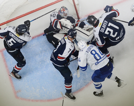 Хоккейный матч возле ворот. Несколько игроков в бело-синих и тёмно-синих формах борются за шайбу. Вратарь в белом обмундировании пытается защитить ворота. Напряжённая игровая ситуация на льду