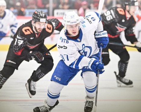 Хоккеисты в белой и черной форме сражаются на льду. Игрок в белом с клюшкой стремится вперед, а другие игроки наблюдают за его маневром. Атмосфера напряженная, зрители на заднем плане