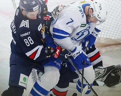 Хоккейный матч: игрок в синей форме сражается за шайбу с игроком в тёмной форме. Эмоции накалены, игра напряжённая. Позади них виден вратарь в красной маске, готовый защитить ворота