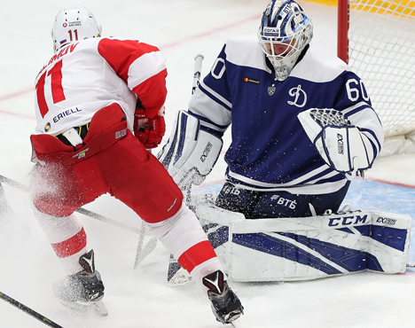 Хоккеист в красной форме атакует, пытаясь забить шайбу, в то время как вратарь в синей форме защищает ворота. Оба игрока напряжены, лёд вокруг ворот раскидан