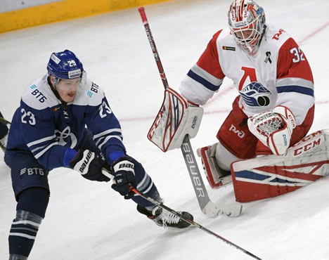 Хоккеист в синей форме атакует у ворот, где вратарь в белой и красной форме готовится отбить шайбу. Динамичная сцена на льду, игроки сосредоточены на игре, держат клюшки, обе команды борются за гол